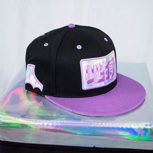 May include: A black and purple baseball cap with a holographic patch on the side and a purple brim. The patch has Japanese characters on it.