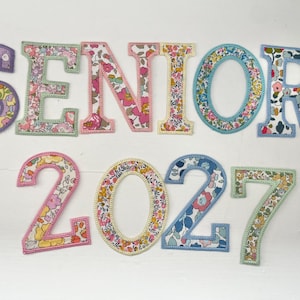 May include: A set of fabric letters and numbers spelling SENIOR 2027. Each character features a unique floral pattern in various colors including pink, blue, yellow, and green, with a stitched border outlining the shape of each individual letter and number.