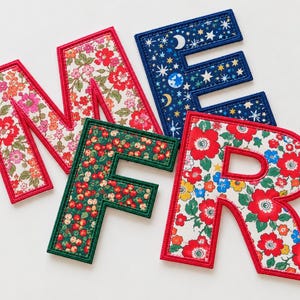 May include: Four fabric letters spelling M, E, F, R. The M and R feature a red, pink, and white floral pattern with red stitching. The E has a dark blue background with white stars, moons, and planets. The F has a green background with red berries.