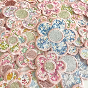 Puede incluir: Primer plano de numerosos parches de tela con forma de flor. Cada parche presenta un borde bordado en rosa y un centro brillante. Los diseños de tela muestran estampados florales en rosas, azules, amarillos y blancos, creando una exhibición colorida.