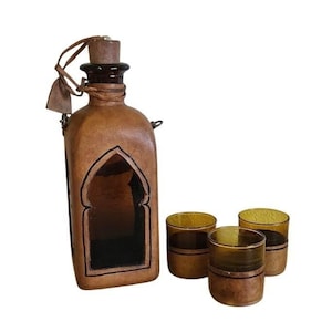 Puede incluir: Una botella de vidrio alta recubierta de cuero color canela con tapón de corcho y correa de cuero, que presenta una ventana recortada en arco. Junto a la botella hay tres vasos de vidrio color ámbar, cada uno con una base de cuero a juego.