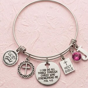 May include: A silver-tone expandable wire bangle bracelet featuring five charms: a round disc engraved with "GOD is Good", a circular cross charm, a round disc with "I CAN DO ALL THINGS THROUGH CHRIST WHO STRENGTHENS ME PHIL 4:13", a rectangular "HOLY BIBLE" charm, a pink crystal, and a letter "J".