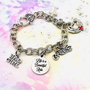 May include: A silver charm bracelet featuring a variety of charms, including a motorcycle, a winged heart, a cross, and a charm that reads "Bad Girl". There is also a round charm inscribed with the words "Life is a Beautiful Ride".