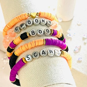 May include: Stack of colorful bracelets with the words "SPOOKY", "BOO", and "SCAR" spelled out in silver letter beads. The bracelets are made of orange, black, purple, and peach-colored disc beads, with gold accents and a heart charm.