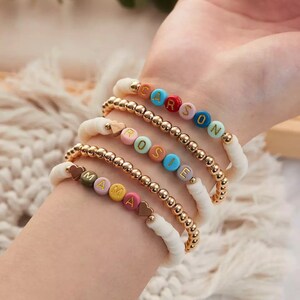 Könnte beinhalten: Ein Set aus fünf bunten Perlenarmbändern mit goldenen Perlen und Herzanhängern. Jedes Armband buchstabiert einen Namen: Mama, Rosie, Carson.