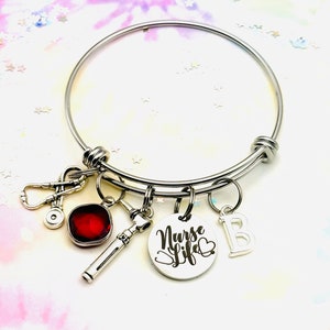 May include: A silver bangle bracelet with charms, including a stethoscope, a red gemstone, a medical tool, and a charm with the text "Nurse Life" and a heart. The bracelet also has a charm with the letter "B".