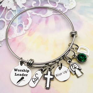 May include: A silver bangle bracelet with multiple charms, including a cross, an angel, a microphone with the text "Worship Leader", a tag with the text "Joy", and a round charm with the text "Rise Up!" The bracelet also features a green gemstone and a charm with the letter "D".