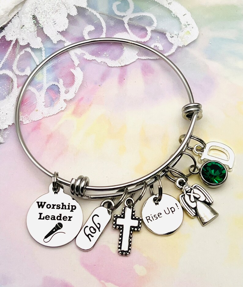 May include: A silver bangle bracelet with multiple charms, including a cross, an angel, a microphone with the text "Worship Leader", a tag with the text "Joy", and a round charm with the text "Rise Up!" The bracelet also features a green gemstone and a charm with the letter "D".