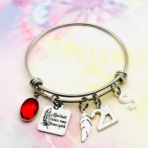 May include: Silver bangle bracelet with charms. The charms include a red gemstone, a feather, a square charm with the text "Kind heart, Fierce mind, Brave spirit", a number 21 charm, and a letter S charm.