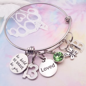 Peut inclure: Un bracelet à breloques en argent avec plusieurs breloques, dont un cristal vert, un papillon, une lettre 'B', le chiffre '13' et un breloque avec le texte 'be bold be brave be you' et 'Loved'.