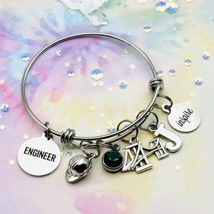 May include: A silver charm bracelet with multiple charms, including a green gemstone, a miniature hard hat, a charm that says "Engineer", a charm that says "Inspire", and a charm that looks like the Golden Gate Bridge.