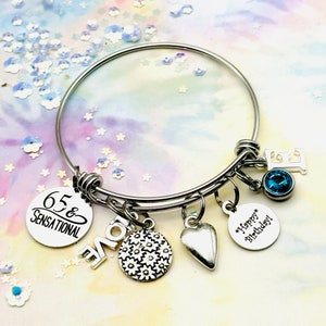 Op de afbeelding: Zilveren armband met meerdere bedels, waaronder een bedel met de tekst "65 & Sensational", een hartbedel, een bedel met een bloemenontwerp, een bedel met de tekst "Happy Birthday" en een blauwe kristallen bedel.