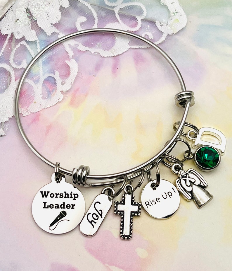 May include: A silver bangle bracelet with multiple charms, including a cross, an angel, a microphone, a tag that says "Worship Leader", a tag that says "Rise Up!" and a tag that says "Joy". The bracelet also has a green gemstone and a charm with the letter "D".