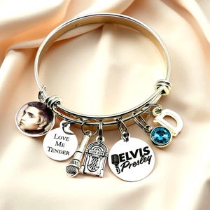 May include: A silver-toned expandable bangle bracelet featuring multiple charms: a circular Elvis Presley portrait, a round disc engraved with Love Me Tender, a microphone charm, a jukebox charm, a disc with Elvis Presley text, a blue crystal, and a letter D.