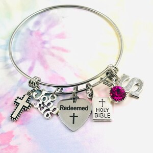 Puede incluir: Un brazalete de plata con dijes que dicen "Juan 3:16", "Redención", "Santa Biblia" y una cruz. El brazalete también tiene un strass rosa y una letra "S" de plata.