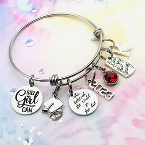 May include: A silver charm bracelet with multiple charms, including a graduation cap, a red gemstone, and charms with the text "This Girl Can", "She believed she could so she did", "Kind is the joy of the journey", and "2023".