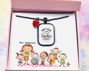 May include: A black necklace with a pendant inscribed with "Most Loved Teacher" and a pencil illustration. A red heart charm is next to the pendant. The necklace is in a pink gift box with a teacher and student drawing.