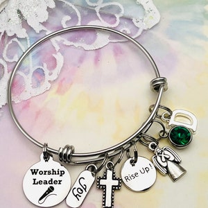 May include: A silver bangle bracelet with multiple charms, including a cross, an angel, a microphone, a tag that says "Worship Leader", a tag that says "Rise Up!" and a tag that says "God". The bracelet also has a green gemstone charm.
