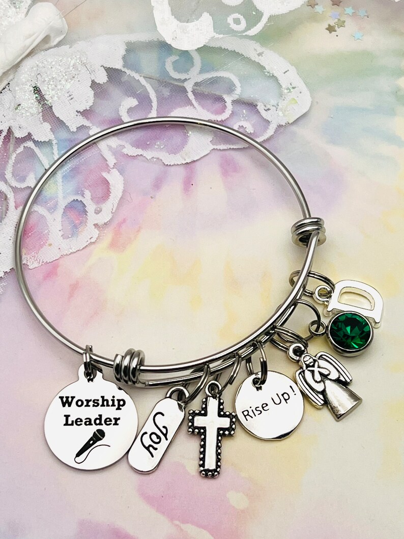 May include: A silver bangle bracelet with multiple charms, including a cross, an angel, a microphone, a tag that says "Worship Leader", a tag that says "Rise Up!" and a tag that says "God". The bracelet also has a green gemstone charm.
