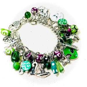Peut inclure: Un bracelet à breloques argenté avec des perles vertes et violettes, des breloques d'extraterrestres, un OVNI, une étoile et le mot "croire".