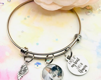 Könnte beinhalten: Ein silbernes, verstellbares Armreif-Armband mit drei Anhängern. Ein Anhänger ist eine silberne Flügel, ein anderer ist ein runder Foto-Anhänger mit einem Katzenkopf und der dritte ist ein runder silberner Anhänger mit dem Text "A piece of my heart lives in Heaven".