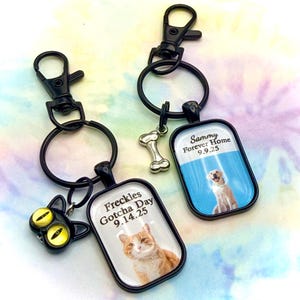 May include: Two rectangular keychains with black metal frames and lobster clasps. One features a cat with text "Freckles Gotcha Day 9.14.25" and a cat head charm. The other shows a dog with text "Sammy Forever Home 9.9.25" and a silver bone charm.