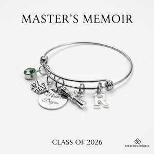 Puede incluir: Una pulsera de brazalete plateada con múltiples dijes. Los dijes incluyen un birrete, un diploma, un disco con "Master's Degree" y la letra "R". La pulsera también tiene una gema verde. El texto en la imagen dice "MASTER'S MEMOIR" y "CLASS OF 2026".