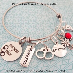 May include: A silver charm bracelet with charms that say "Partners in Crime", "Fearless", "Strength", and a handcuff charm. The bracelet also has a red gemstone and a flower charm.