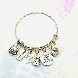 May include: Silver charm bracelet with multiple charms, including a cake, the number 21, a dress, a flower, a crystal, and a charm that says "Follow Your Yellow Brick Road".