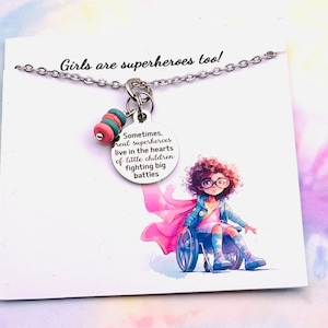 May include: A silver necklace with a charm that says "Sometimes, real superheroes live in the hearts of little children fighting big battles". The charm is attached to a pink and teal beaded chain. The necklace is displayed on a white card with a watercolor illustration of a girl in a wheelchair wearing a pink cape and glasses.