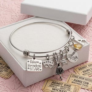 May include: A silver-toned metal bangle bracelet featuring multiple charms: a square tag reading Future Broadway STAR, a theatrical mask, a dancer silhouette, a star, a round tag reading Dream Big, a yellow teardrop gem, and the letter A.