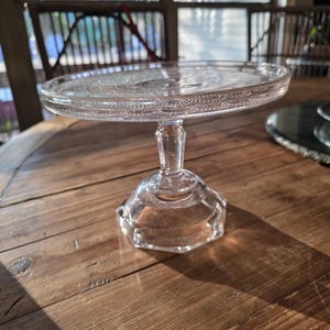 May include: A clear glass cake stand with a round top and a pedestal base. The top has a decorative border. The stand is on a wooden table. The stand is approximately 10 inches in diameter.