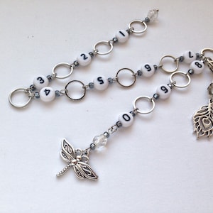 Könnte beinhalten: Eine silberne Kette mit weißen Perlen, die von 0 bis 9 nummeriert sind, und einem Libellen-Anhänger. Die Kette hat auch einen silbernen Feder-Anhänger.