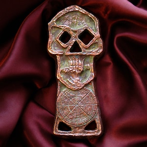 May include: A bronze-coloured, skull-shaped decorative item with a compass design at the base. The skull has diamond-shaped eye sockets and a textured surface. The item is displayed on a deep red, satin-like fabric.