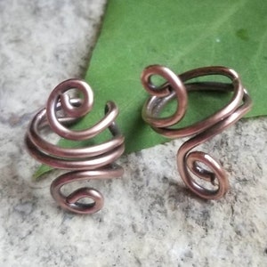 May include: Two copper wire ear cuffs, each with a unique swirling design. The cuffs are shown on a textured surface.