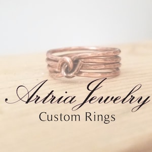 May include: A close-up of a copper ring with a knot design. The ring is made of multiple bands of hammered copper. The text "Artria Jewelry Custom Rings" is visible below the ring.