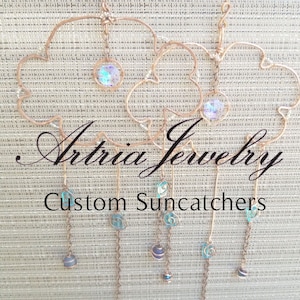 May include: Two copper wire cloud suncatchers with iridescent glass beads and blue glass beads hanging from the bottom. The suncatchers are suspended from a copper wire loop. The text "Artria Jewelry Custom Suncatchers" is visible below the suncatchers.