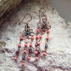 Puede incluir: Pendientes de alambre de cobre con un diseño delicado e intrincado. Los pendientes presentan una pequeña piedra preciosa iridiscente en el centro del diseño y cuelgan con múltiples hebras de cuentas de color coral.