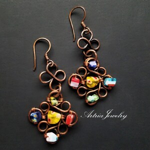 May include: A pair of copper wire earrings with colorful glass beads. The earrings are shaped like a clover with a loop at the top for hanging. The beads are red, yellow, blue, green, and white.