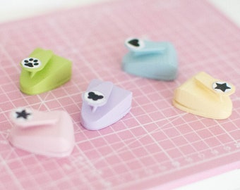 May include: Five different colored paper punch tools with various shapes, including a star, a paw print, and a cloud, on a pink cutting mat.