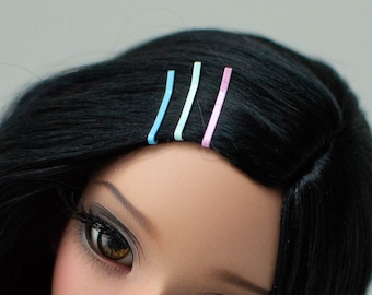 May include: A doll with long black hair, wearing a white shirt with a pink and purple pattern. The doll has brown eyes and is wearing pink, blue, and white hair clips.