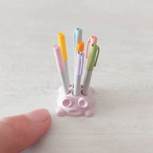 May include: A pink plastic pen holder shaped like a cartoon face with six holes for pens. The holder is filled with six colourful pens, including yellow, orange, green, blue, and purple.