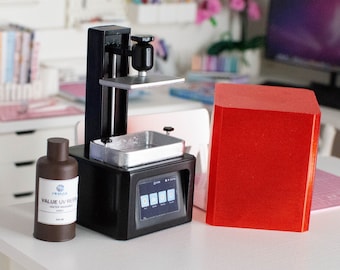 May include: A miniature black 3D printer with a silver tray and a brown bottle labeled "VALUE UV RESIN" sits on a white table. A red rectangular object is next to the printer. The background shows a craft room with colorful supplies.