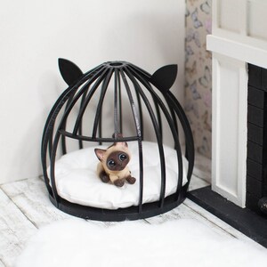 May include: A black wire cat bed with a white cushion and a small Siamese cat figurine inside. The bed is in front of a white fireplace with a black brick surround and a small fire inside.