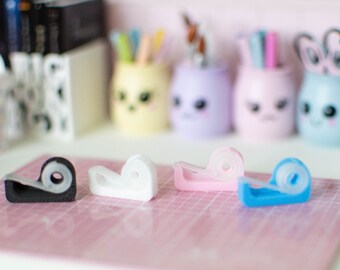 May include: Four miniature tape dispensers in black, white, pink, and blue. The dispensers are sitting on a pink cutting mat.