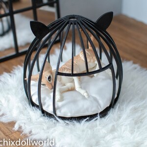 May include: A black cat bed shaped like a cage with a white cushion inside. A tan cat doll is lying inside the bed.