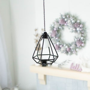 May include: A black metal cage pendant light with a white light bulb hanging from a chain. The light is hanging in front of a white brick fireplace with a wreath on the wall behind it.