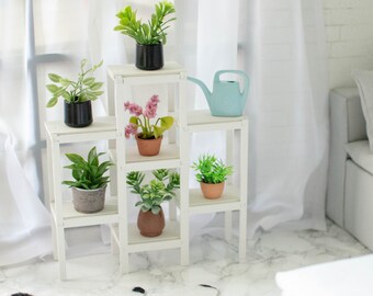 Puede incluir: Estantería blanca de cinco niveles, con plantas en macetas y una regadera azul. La estantería está contra una pared blanca con un sofá blanco en el fondo.