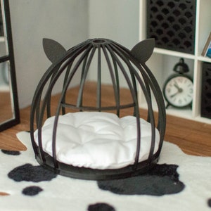 May include: Black cat bed with a white cushion inside. The bed is shaped like a dome with cat ears on top. The bed is on a black and white cowhide rug in front of a white bookshelf with black shelves.