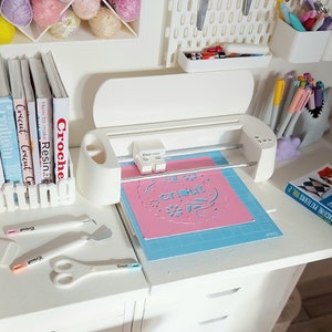 May include: A white crafting machine with a pink mat and a blue cutting mat. The machine is on a white desk with a white drawer. There are several books about crafting on the desk, including books about crochet, knitting, and resin.
